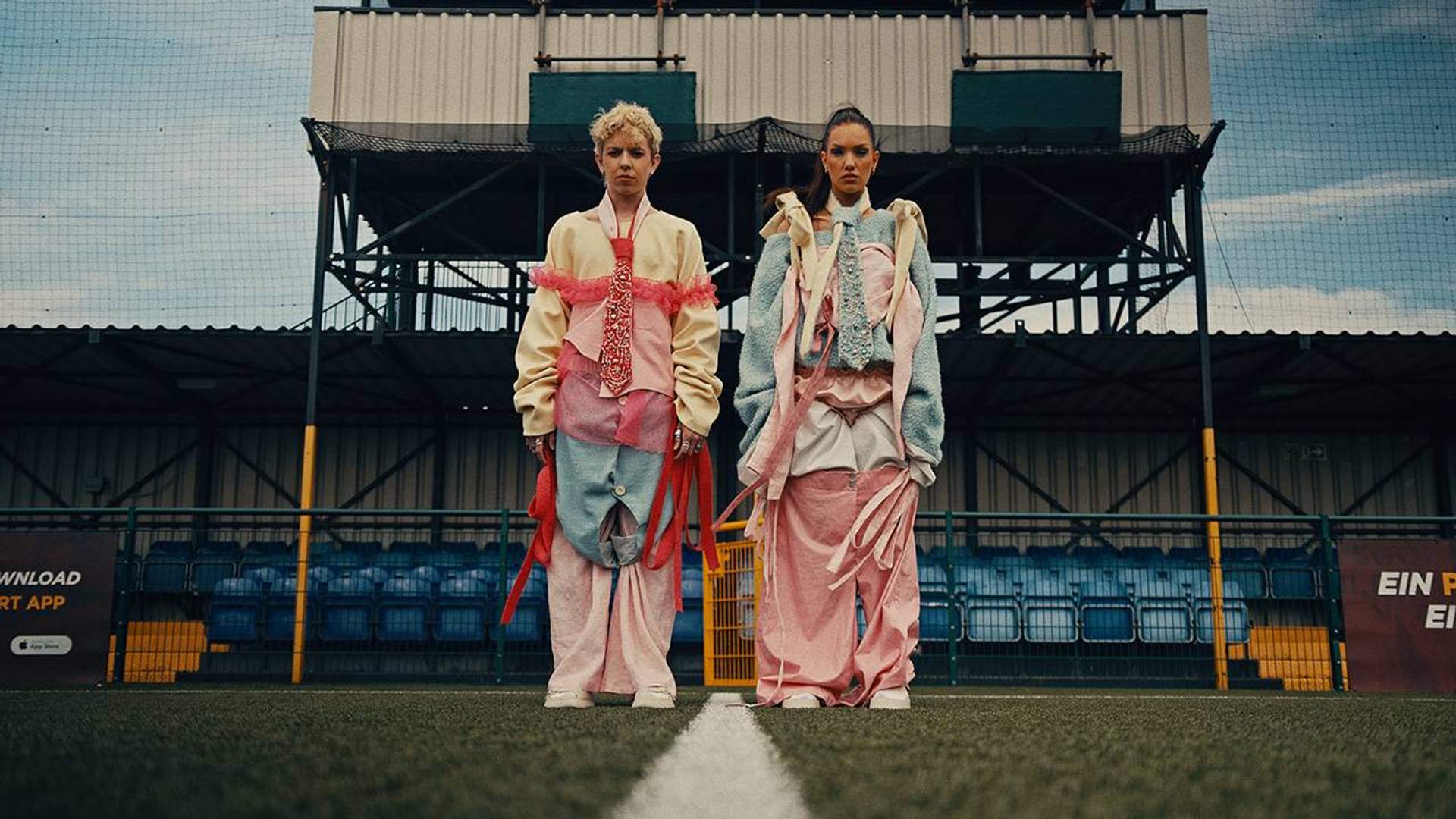 Two people dressed in elaborate colourful outfits pose on a football pitch.