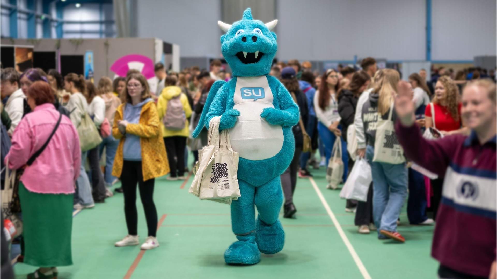 The Students' Union mascot, a light blue and white dragon, holding a tote bag in the middle of a busy Freshers' Fayre