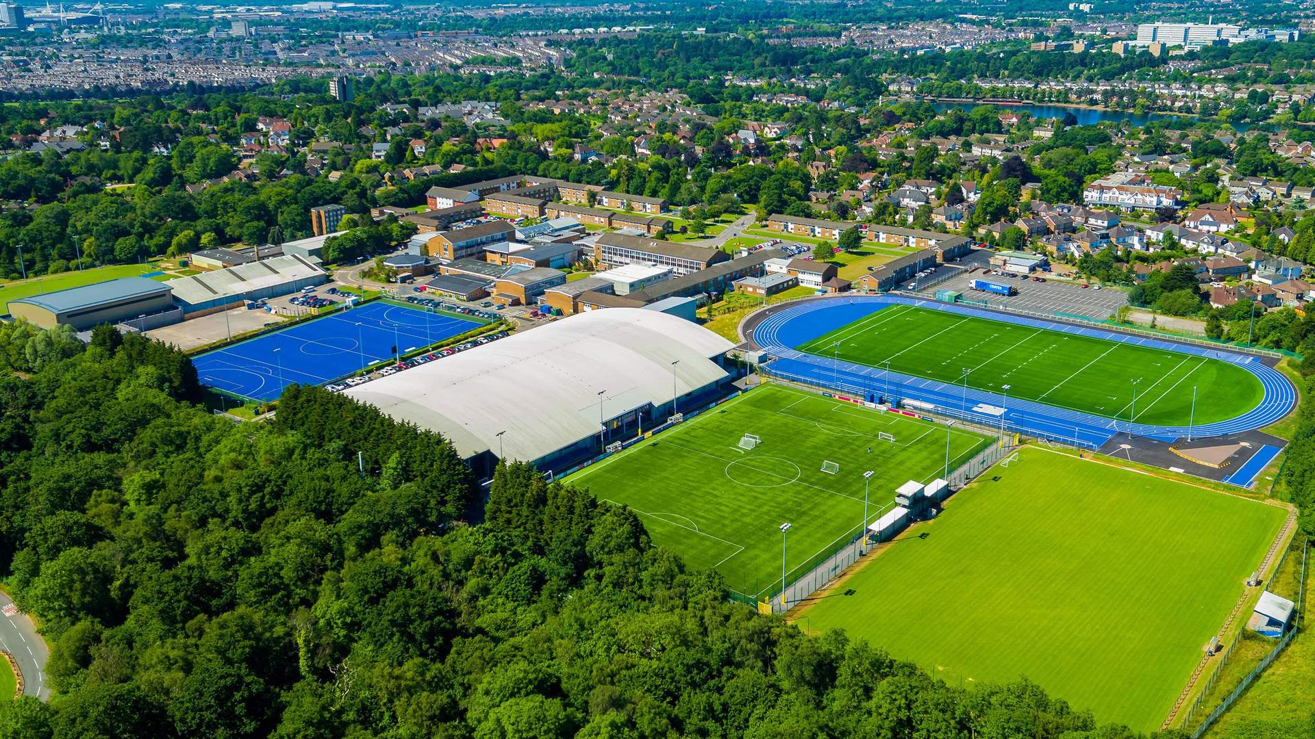 Aerial view of a sports complex featuring a large indoor sports facility, blue athletics track, multiple soccer fields, lush green trees in the foreground, and nearby residential buildings in the background.