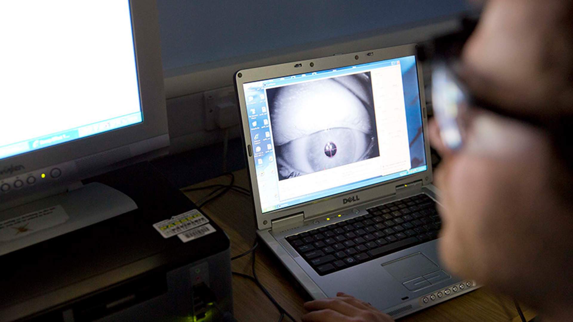A close-up of a person working on a Dell laptop displaying an eye-tracking software interface. The screen shows a magnified image of an eye being analyzed. A second computer monitor and other lab equipment are visible in the background.