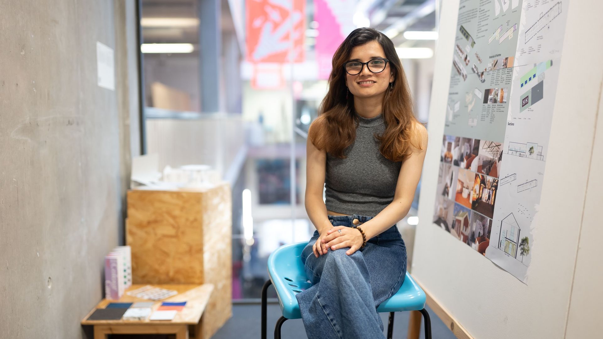 A young woman with long brown hair smiles while sat on a stool. Beside her are images and designs pinned to a whiteboard.