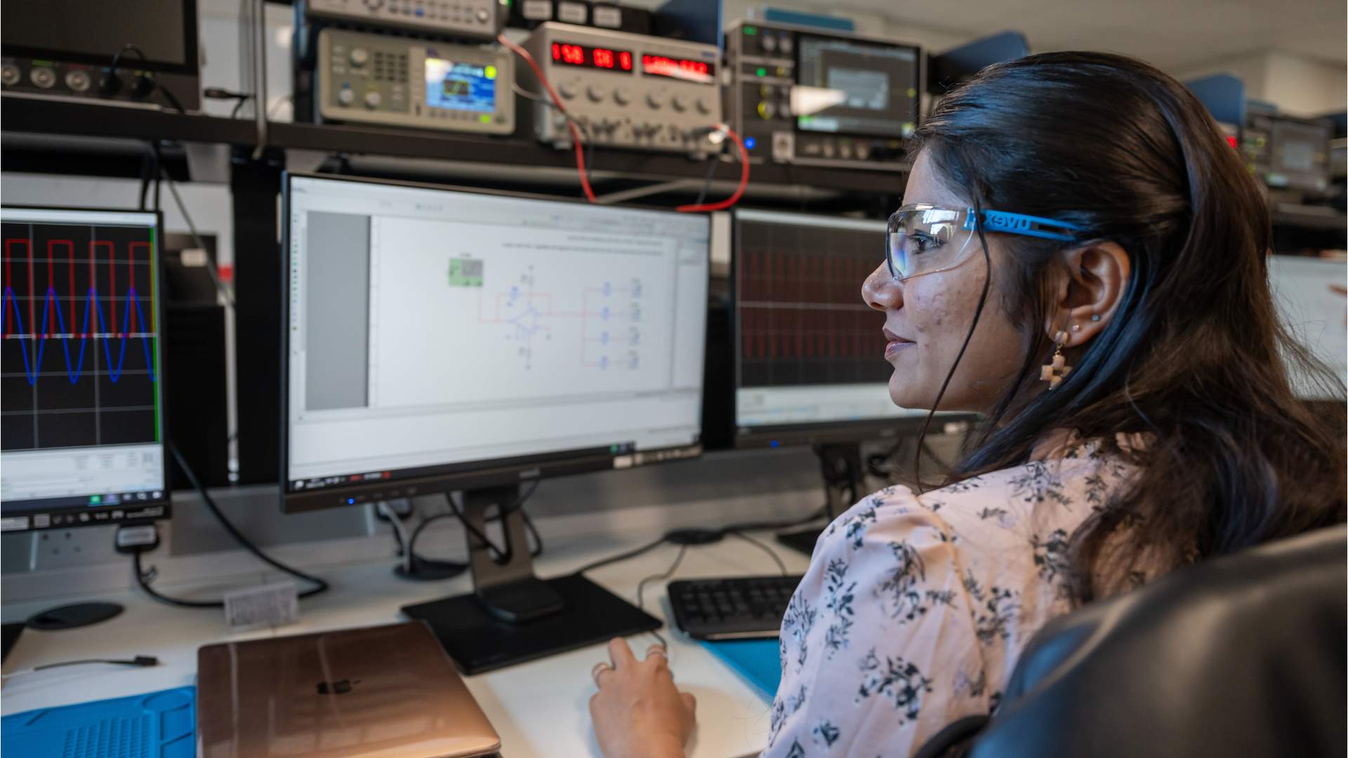 A person with blond hair, wearing a blue patterned shirt, is focused on working with a small robot or electronic device using a screwdriver at a desk in a lab or office setting. Various electronic equipment is visible in the background.