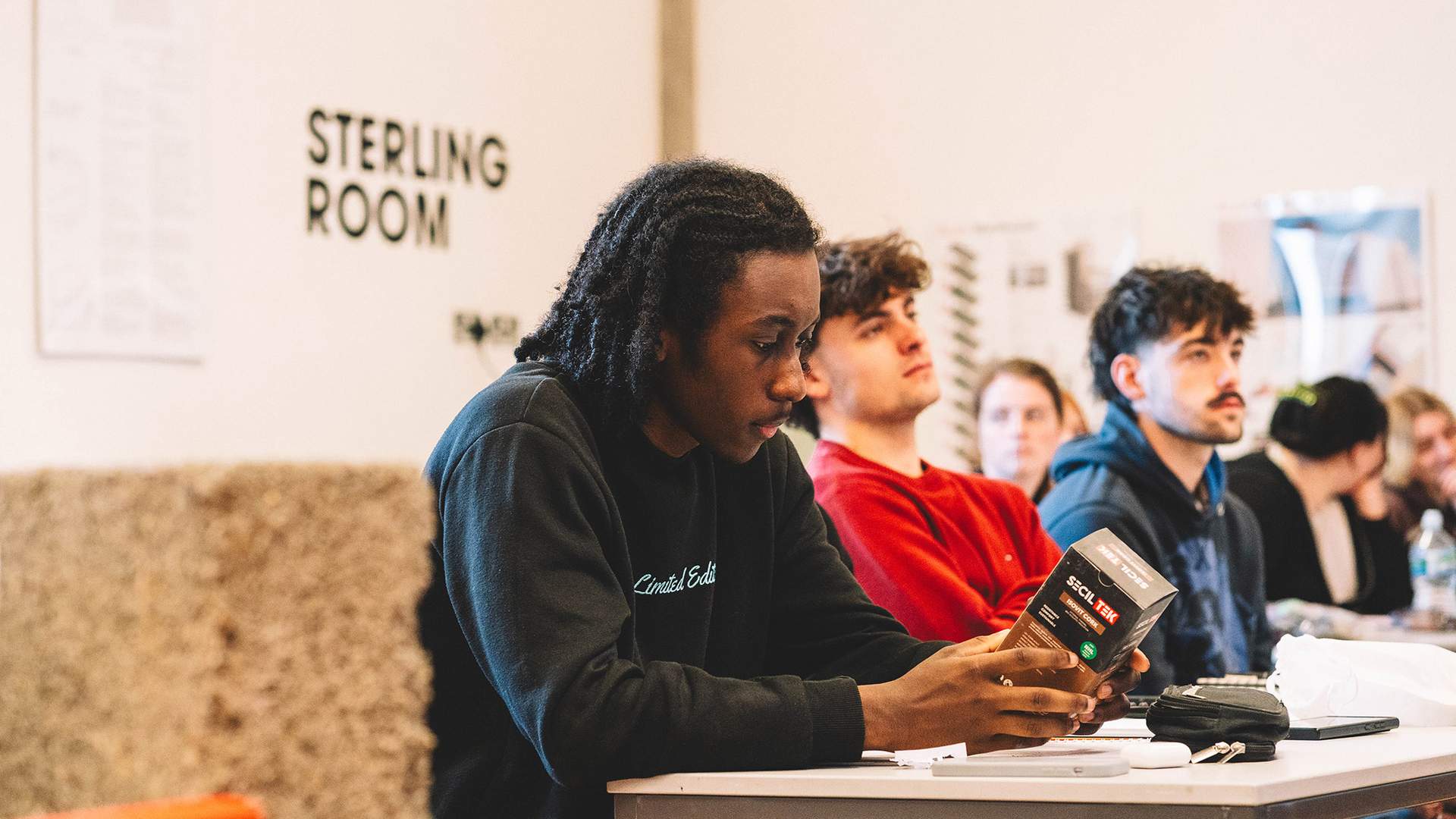 A group of diverse individuals seated around a table in a classroom setting, engaged in discussion and collaboration.