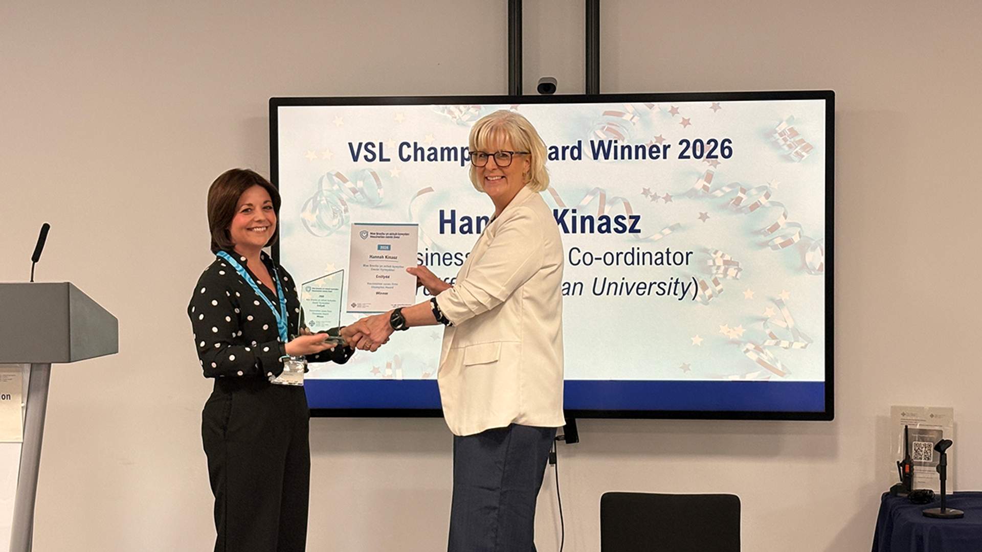 Hannah Kinasz recieving an award from a collegue in front of large monitor