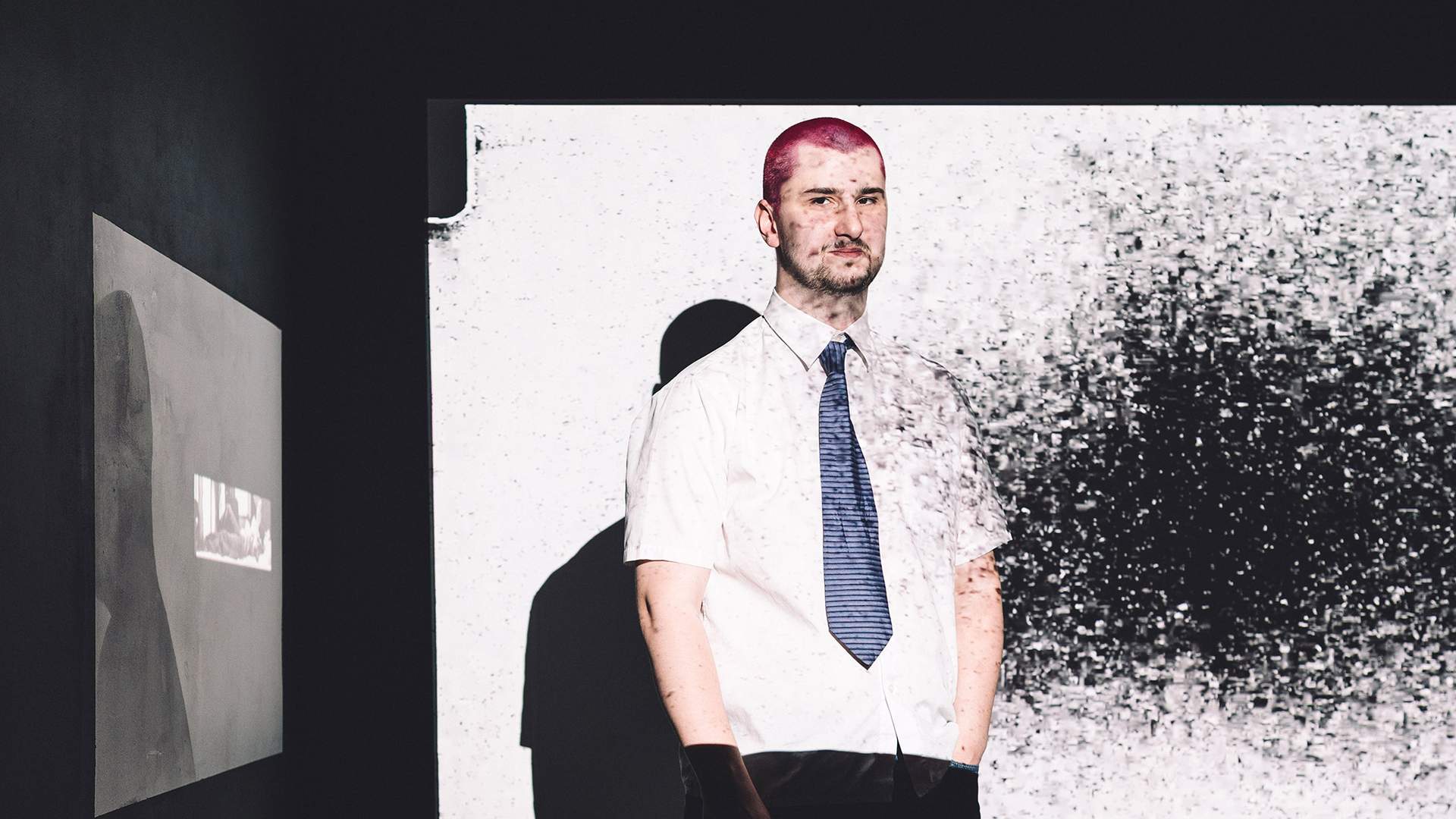 A man with pink hair stands in front of a large screen, engaging with the displayed content.