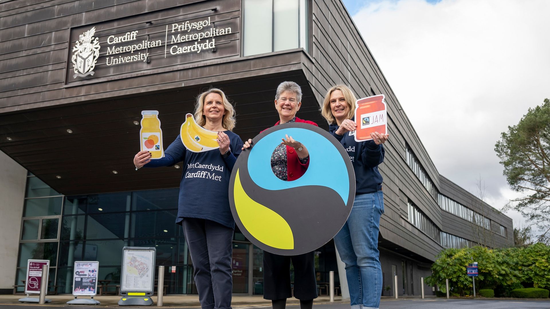Three members of Cardiff Met staff hold Fairtrade information posters outside the Cardiff School of Managament