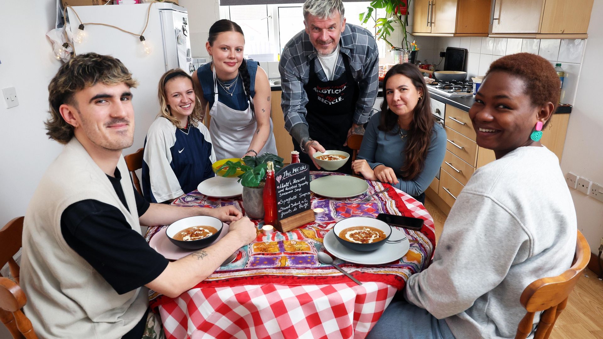 A group of staff and students sit and stand around a decorated kitchen table featuring a spread of various foods