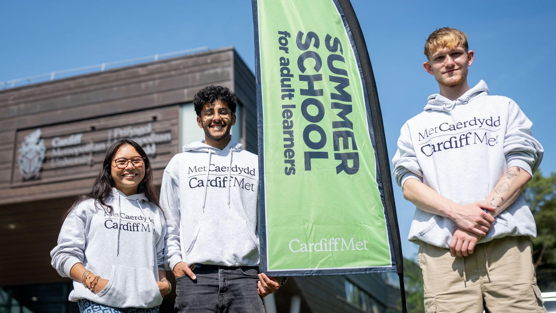 Three young adults in Cardiff Met hoodies stand beside a Cardiff Met Summer School banner