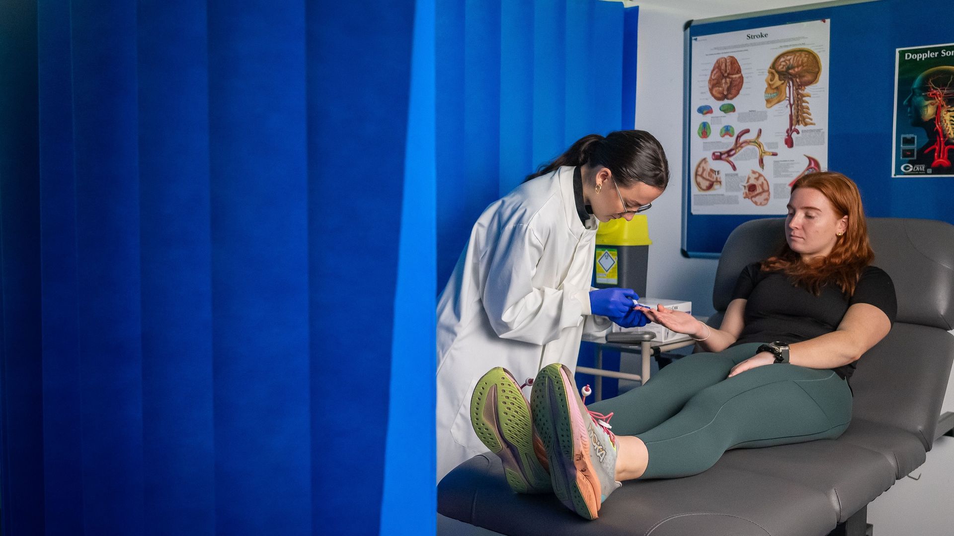 A person in a white lab coat takes a blood sample from the finger of a patient, sat on an examination bed. They are surrounded by a blue floor-to-ceiling curtain.