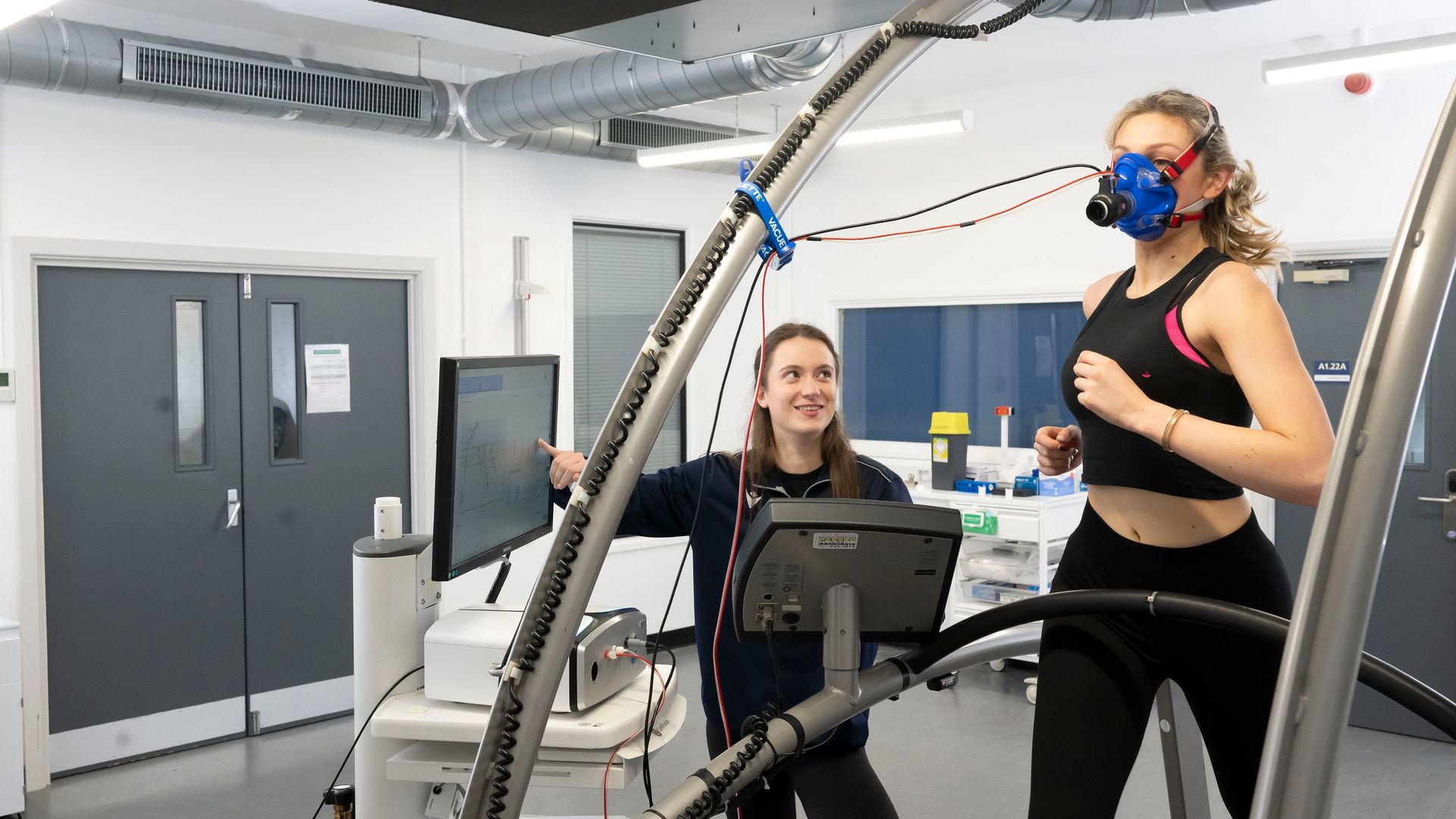 A young woman running on a treadmill is being monitored by another individual pointing at a screen during research