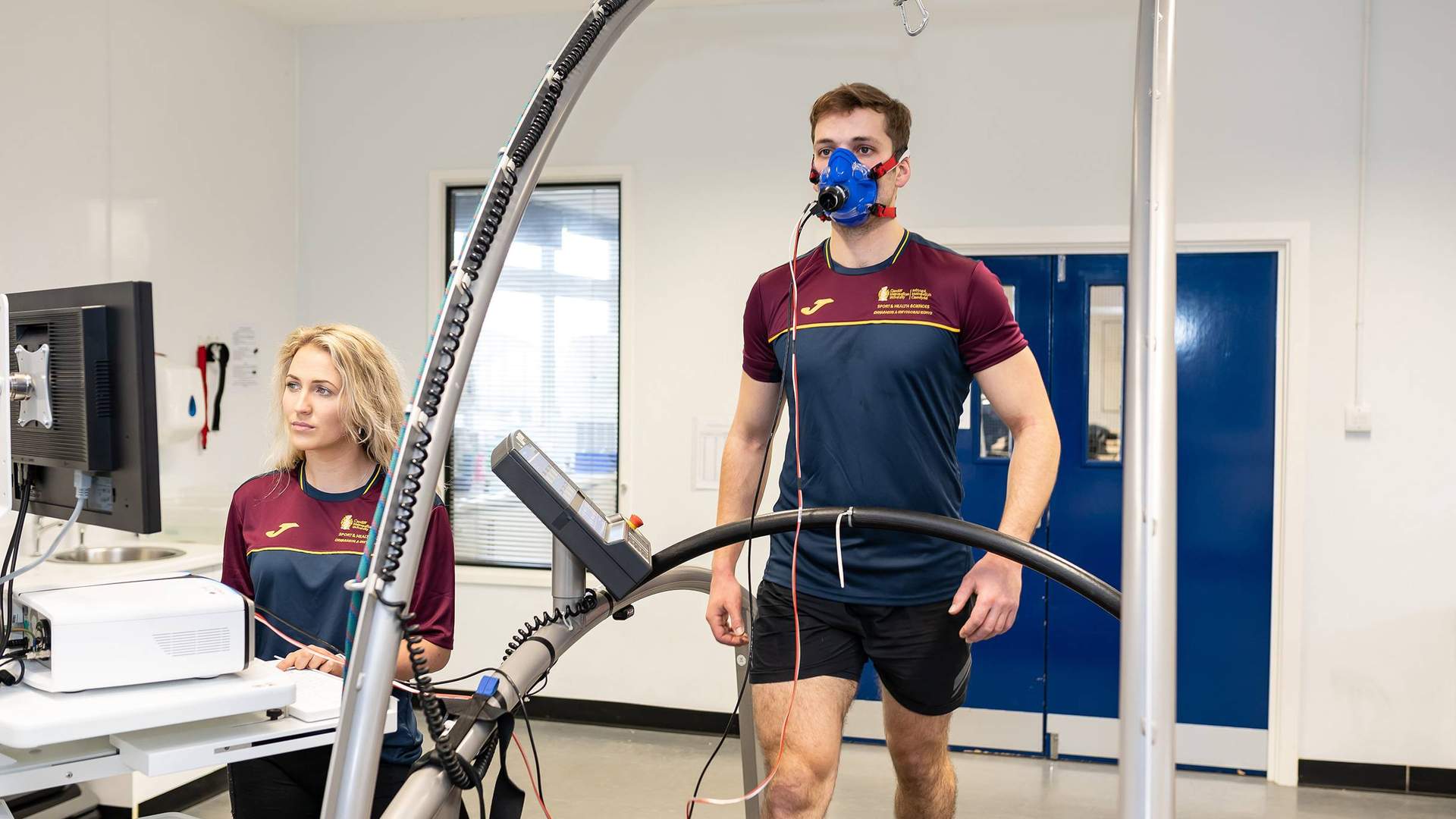 A man is walking on a treadmill wearing a blue mask for a fitness test. A woman stands nearby, operating a computer connected to the equipment. Both are in a lab setting wearing maroon and navy sports attire.