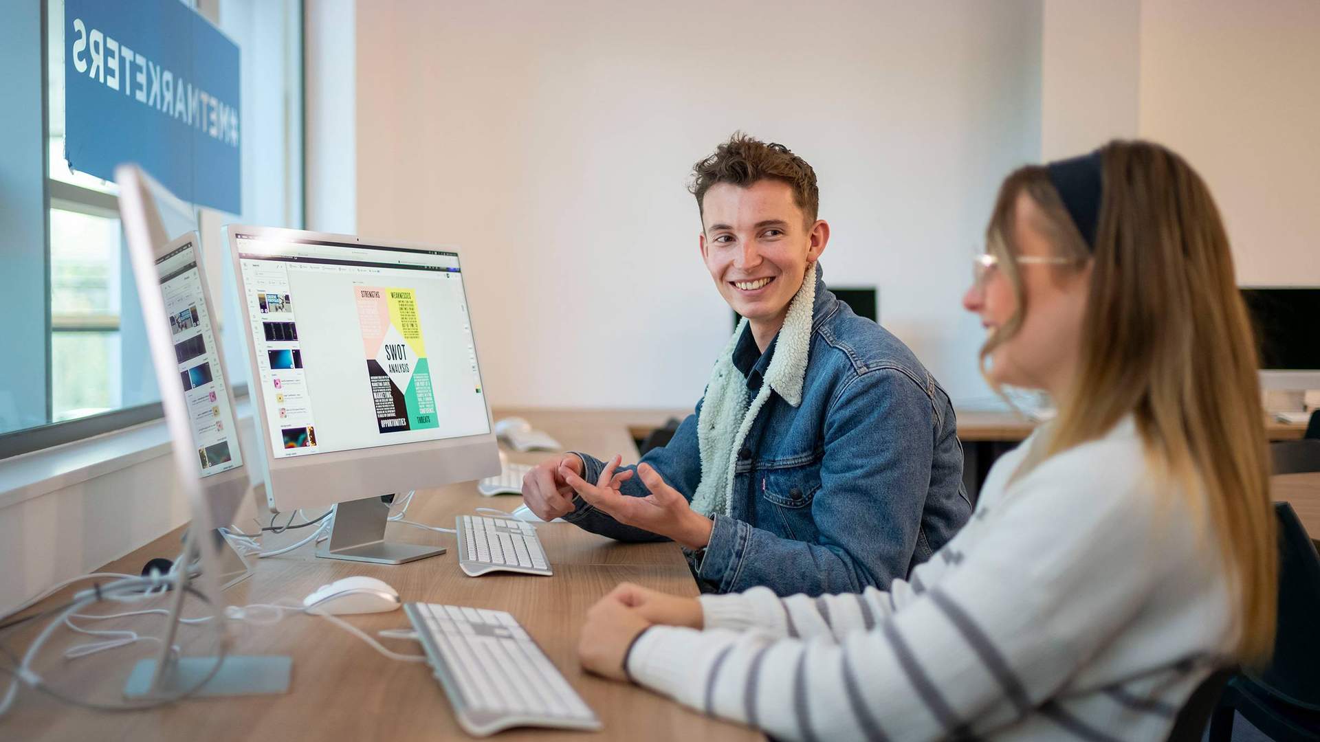 A desk with two people working on computers. One person is talking to the other.