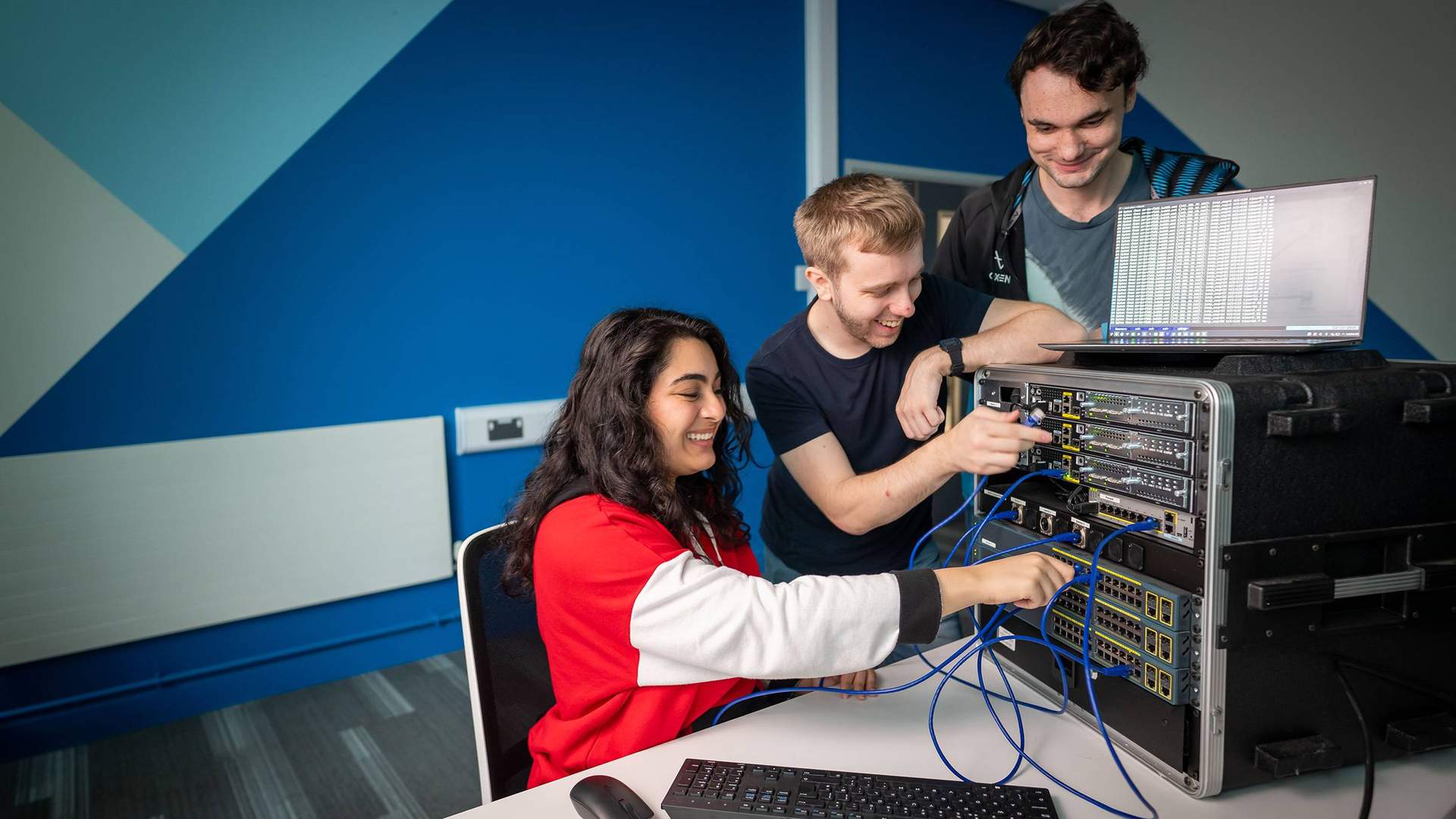 Three students work together, connecting cables to a network switch.