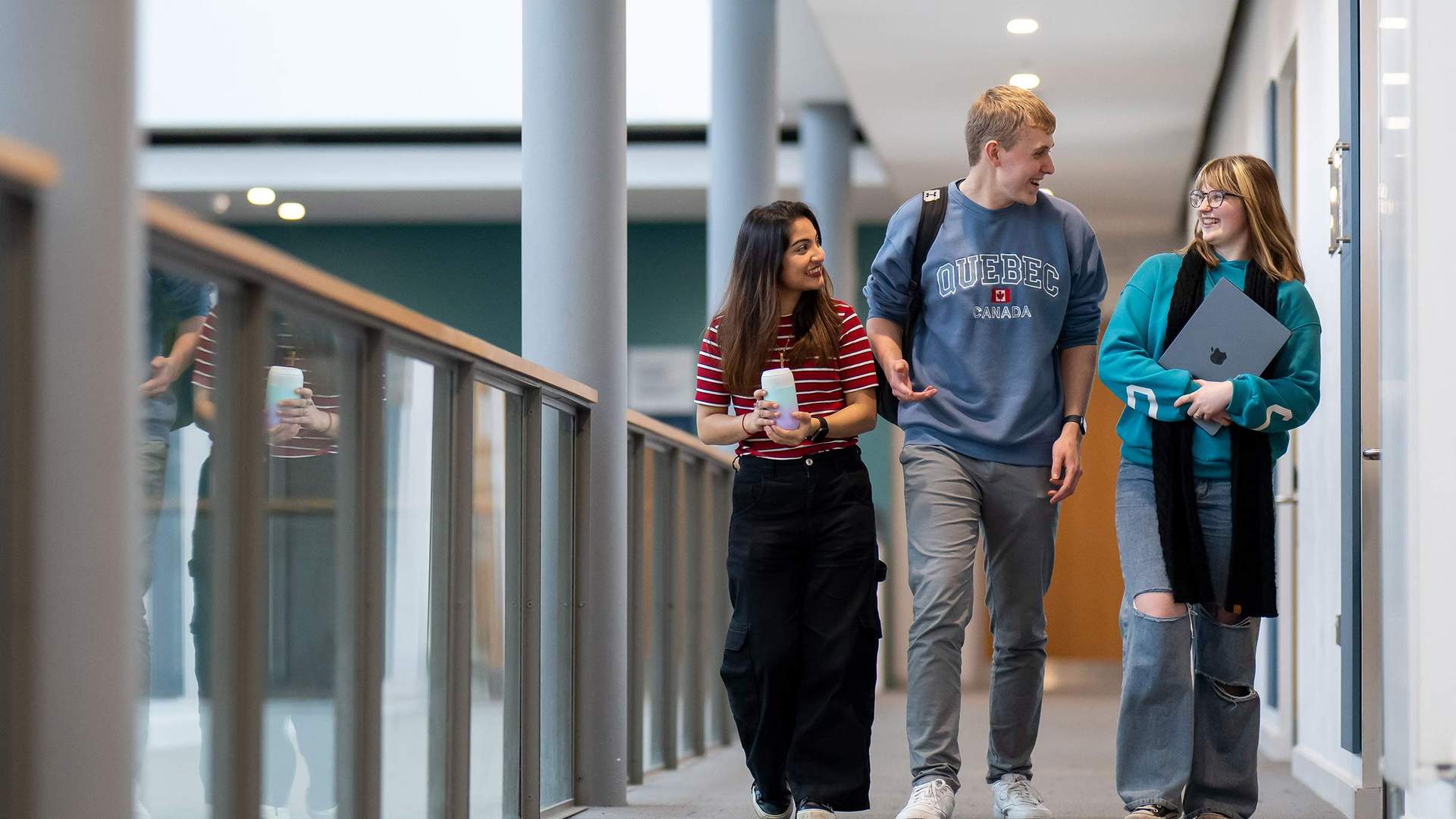 Three young people walk together on a balcony. One is holding a laptop computer.