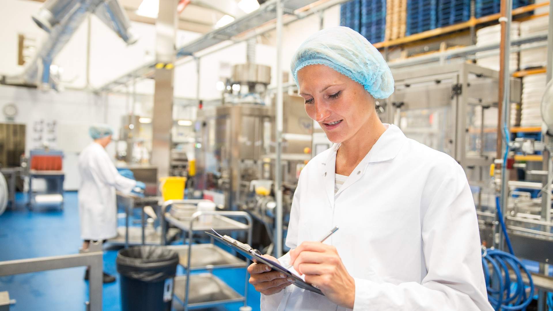 A woman in a white lab coat and hairnet writes on a clipboard in a factory