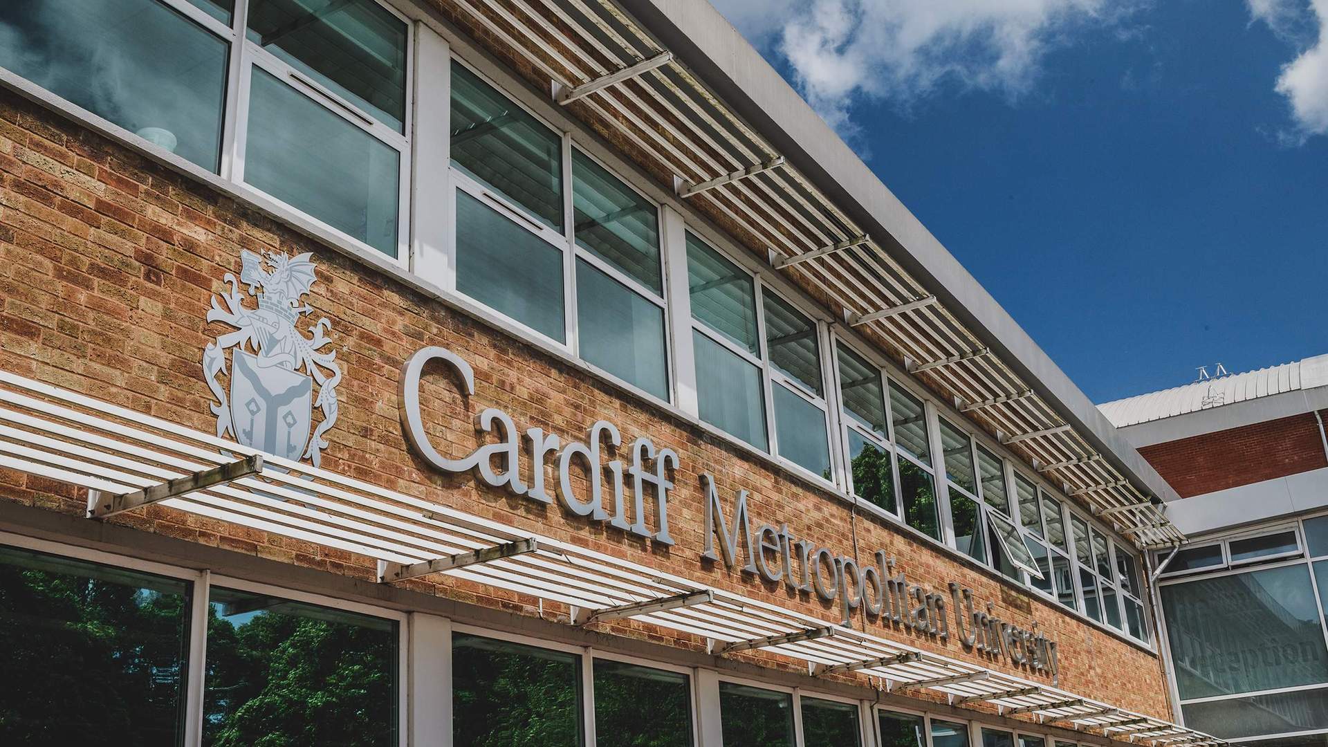 A building with large windows featuring the sign Cardiff Metropolitan University on a brick wall. A blue sky with clouds is visible above.