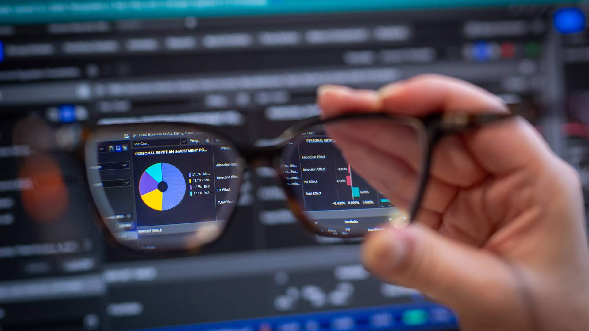 A computer screen displaying stock market data and graphs, viewed through a pair of glasses held in someone's hand.