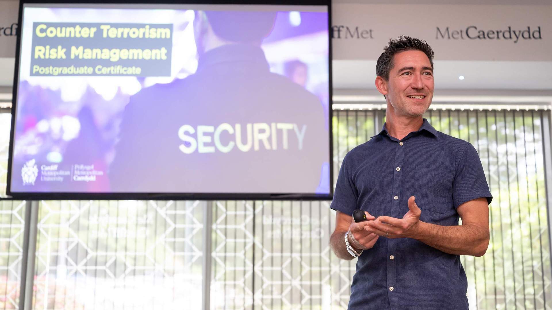 A man stands in front of a screen giving a presentation. The screen shows a person wearing a shirt with SECURITY printed on the back.