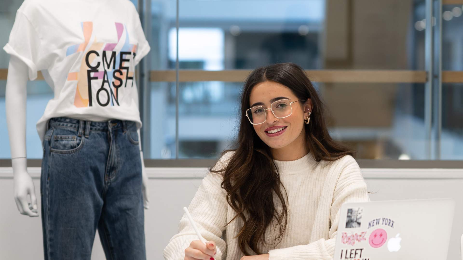 A woman with long hair and glasses sits at a desk smiling, using a stylus on a tablet. She is in front of a laptop with various stickers. Next to her, a mannequin displays a white T-shirt with colorful text. The setting is a modern office.