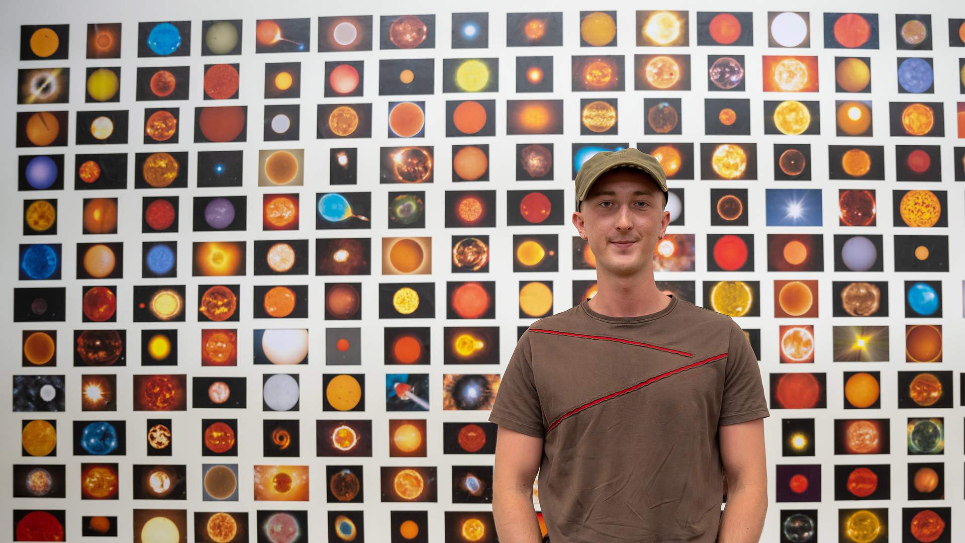 A person wearing a cap stands in front of a large wall collage featuring numerous colorful images of celestial bodies, like stars and planets. The collage is composed of small, square pictures arranged in a grid pattern.