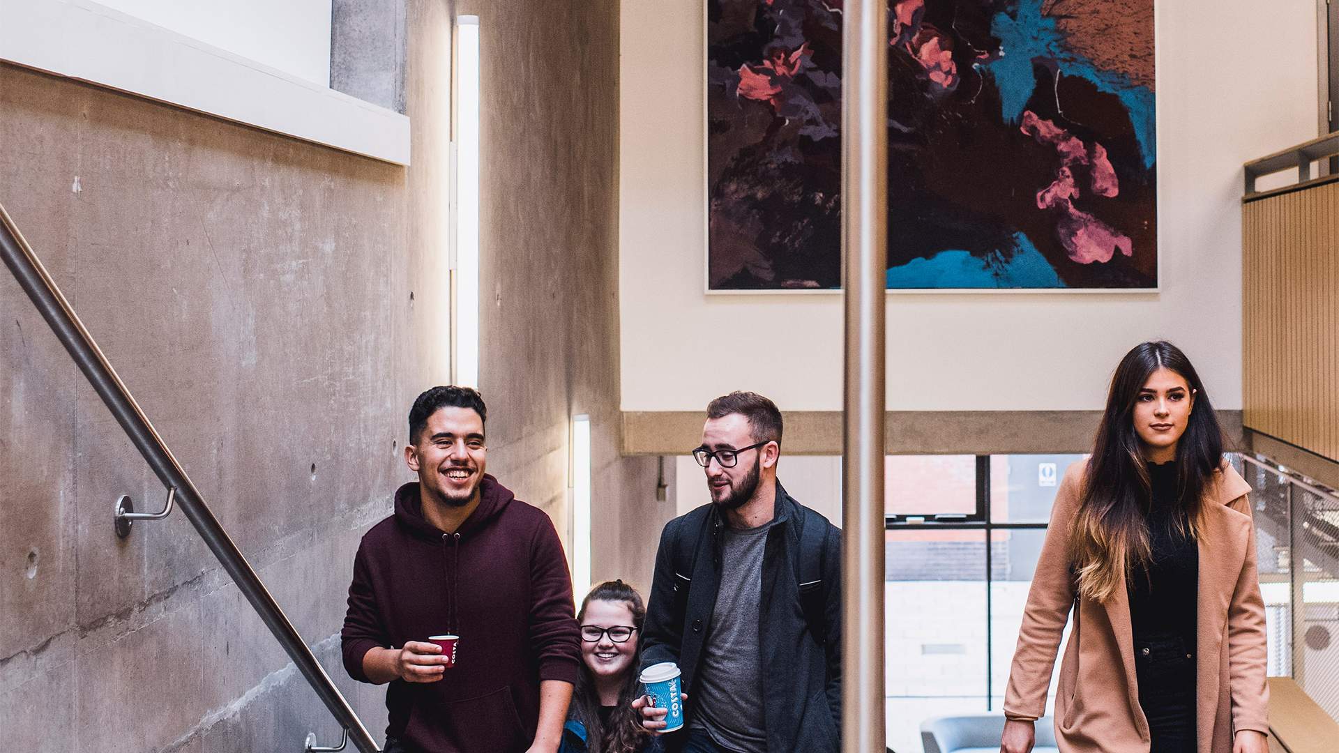 Four people are walking up a staircase, carrying coffee cups. They appear to be engaged in conversation and smiling. The setting is modern with concrete walls and a colorful abstract painting on the upper wall.