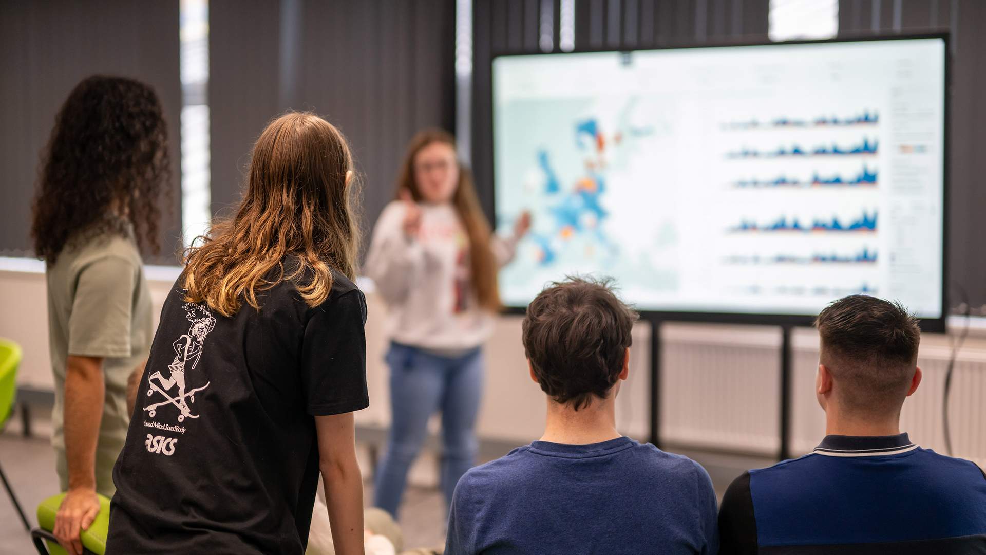 Four people are seated and watching a presentation on data displayed on a large screen, while one person stands in front of the screen explaining the content. The room has a professional setting with green chairs.