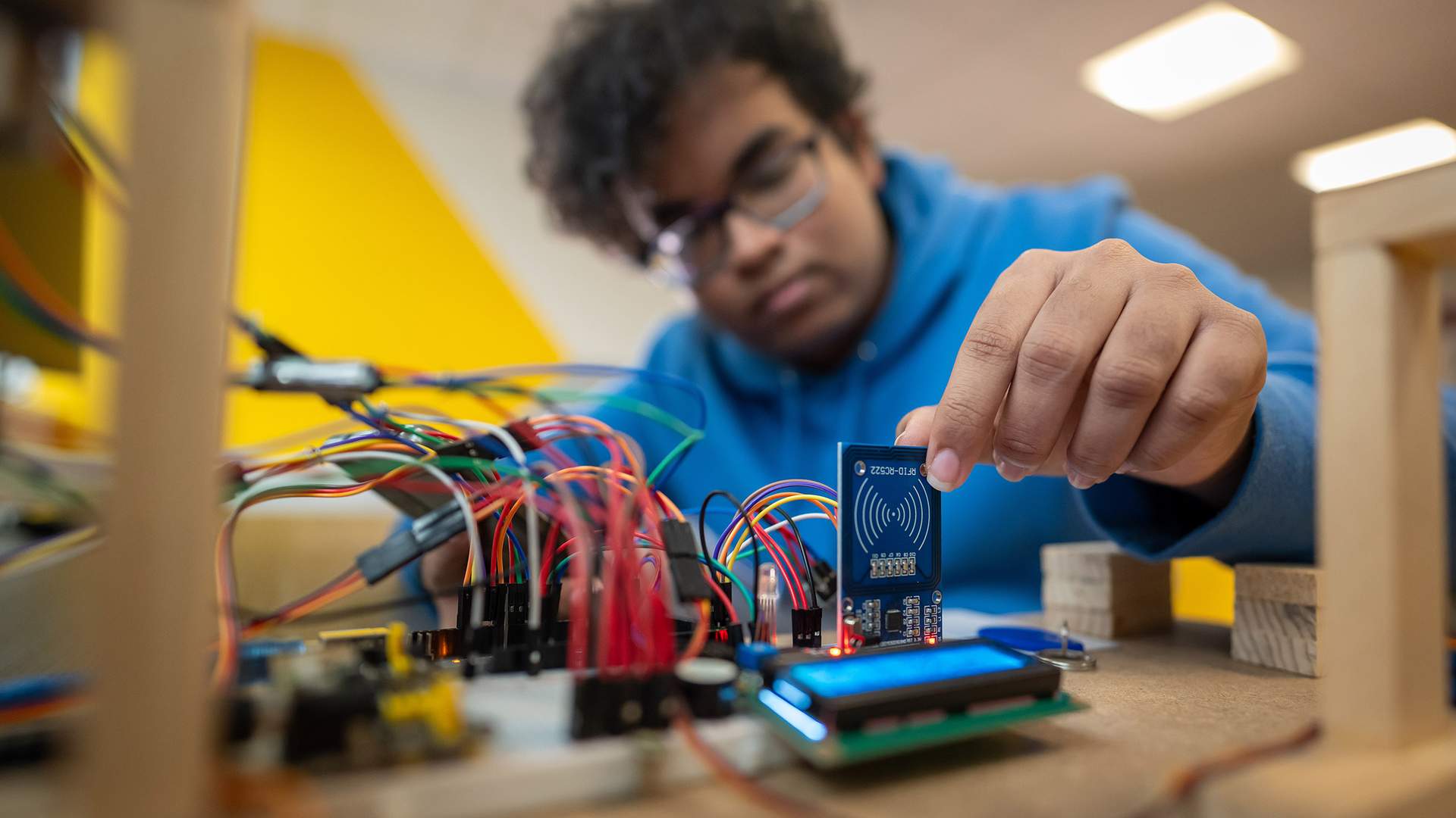 A person in a blue hoodie works with electronic components and circuit boards, placing a small blue board among connecting wires on a table. The background is blurred and the lighting is bright.
