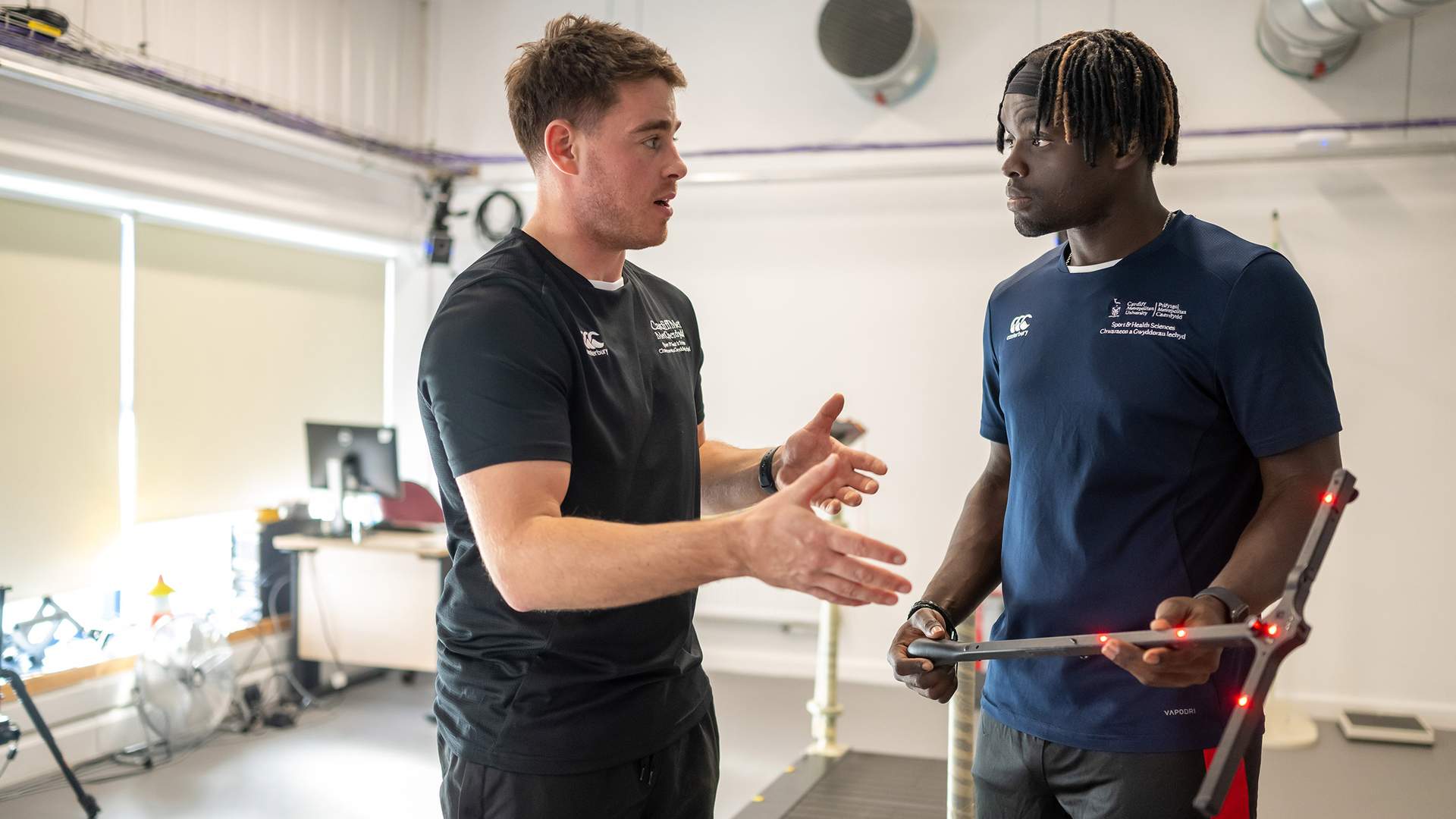 Two men are engaging in conversation in a lab setting. One is gesturing with his hands, while the other holds a device with sensors. Both are wearing athletic gear. A treadmill and office equipment are visible in the background.