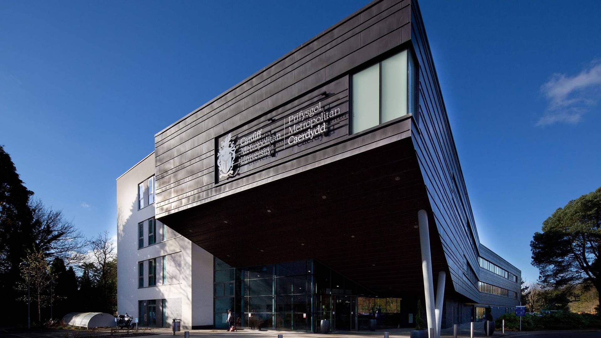 Modern building with angular architecture, featuring a large sign that reads Prifysgol Metropolitan Caerdydd. The structure has a mix of dark panels and white walls, with glass windows. Trees and a blue sky are visible in the background.