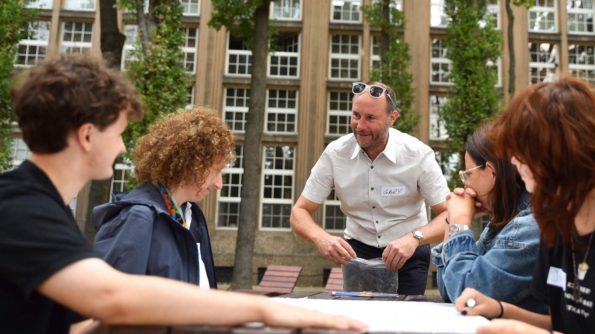 Four people sit at a wooden table outdoors. A man wearing a name badge labelled Gary stands next to them.