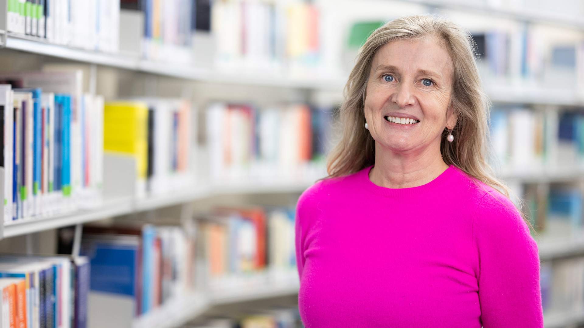 A woman in a bright pink sweater smiles while standing in a library aisle. Shelves filled with various books are visible in the background, creating a scholarly atmosphere.