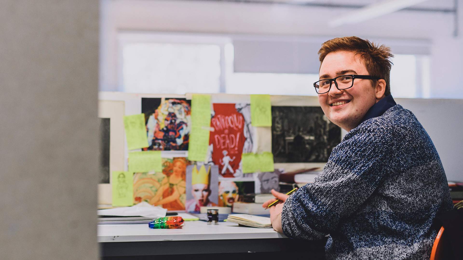A person with short hair and glasses smiles while sitting at a desk covered in colorful images and sticky notes. They hold a pen and notebook, looking towards the camera in a bright, open office space.
