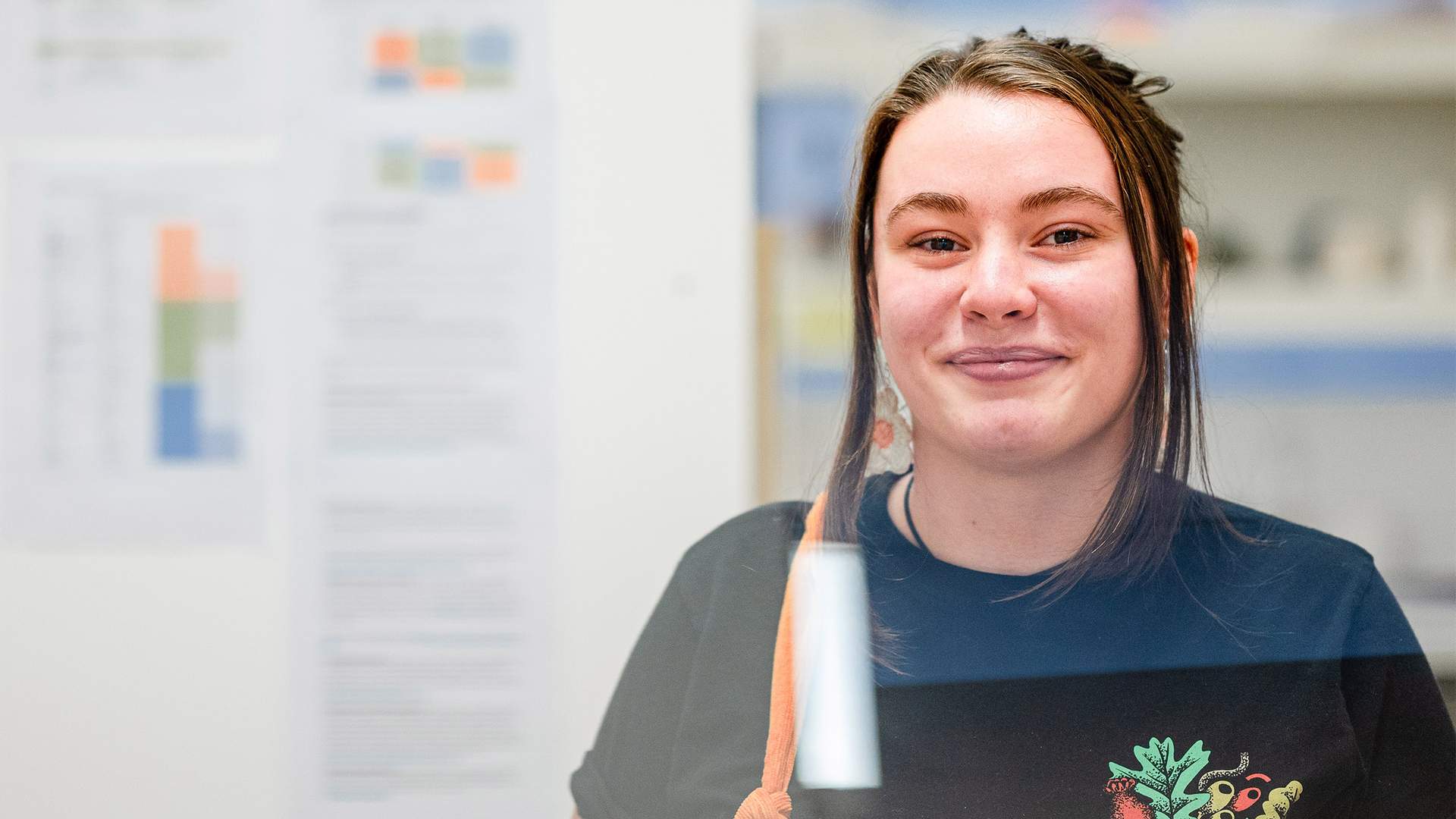 A young woman with long brown hair smiles while standing indoors. She wears a black shirt with floral embroidery. Behind her are blurred documents pinned to a whiteboard.