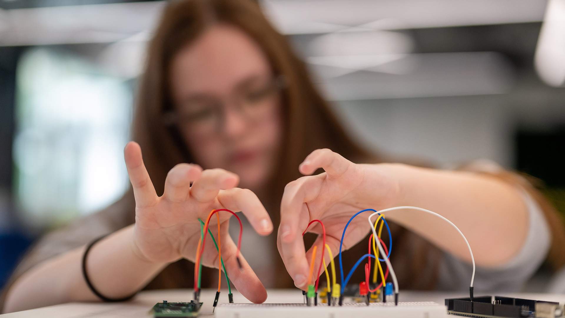 A person with long hair and glasses is working with a breadboard and wires, connecting electronic components. The background is blurred, focusing on the hands and the breadboard.