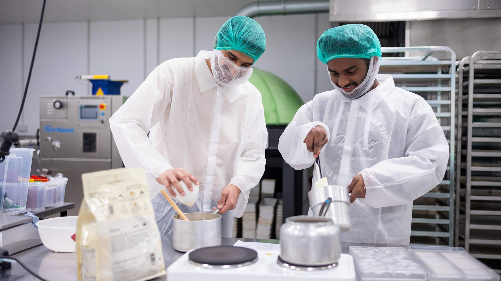 Two people in protective clothing and hairnets work in an industrial kitchen. One person holds a pot while the other pours ingredients into it. Stainless steel racks, equipment, and containers surround them.