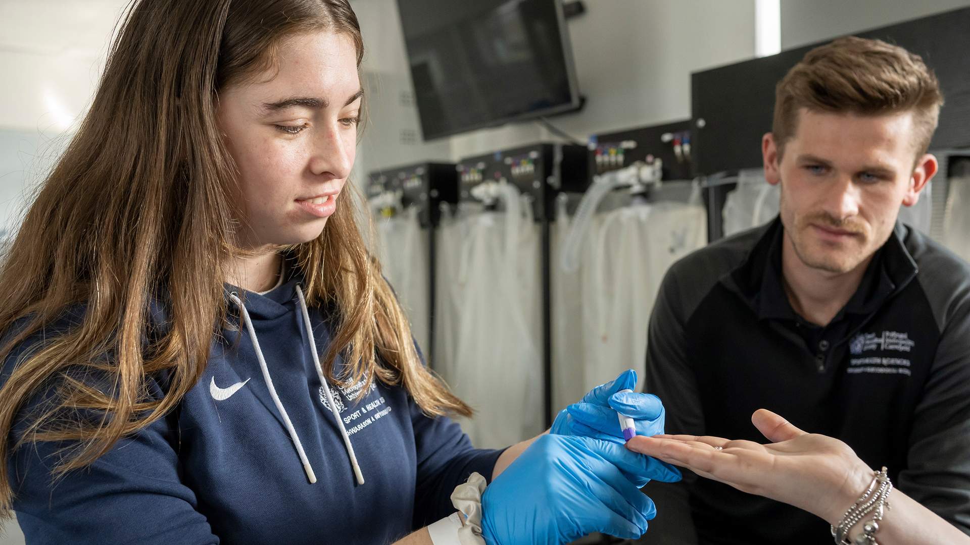 A woman in a blue hoodie and blue gloves is examining the hand of another person. A man in a black shirt observes the process. They are in a room with medical equipment visible in the background.
