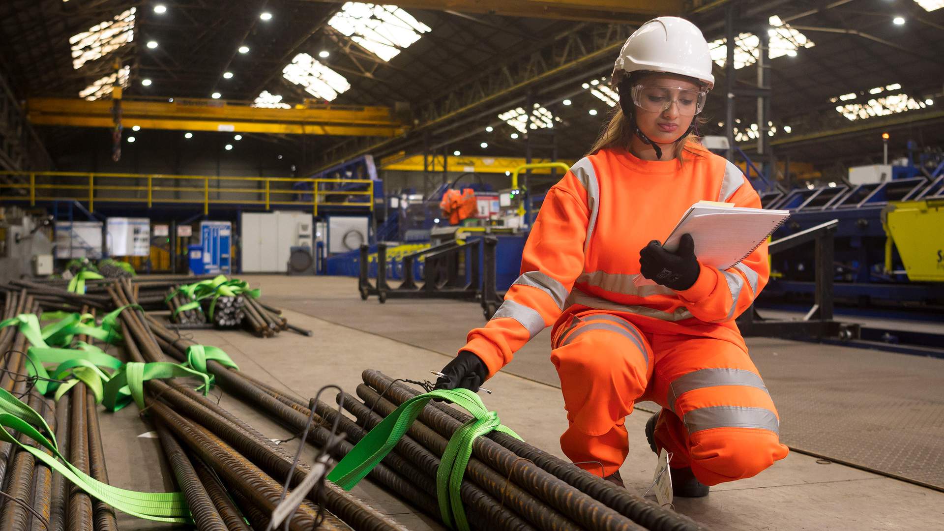 A worker in high-visibility orange gear and a hard hat inspects steel rods tied with green straps in a factory setting. She holds a clipboard, checking the materials under bright overhead lighting.