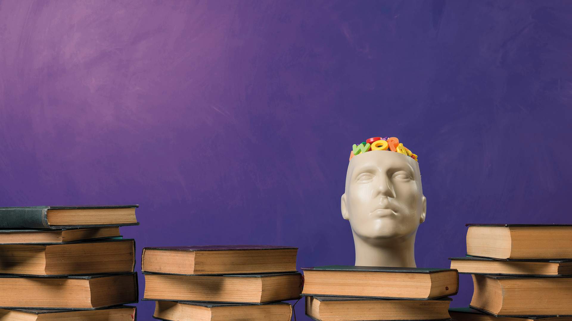 A mannequin head with colorful toy letters inside sits on a table surrounded by stacks of books against a purple background.