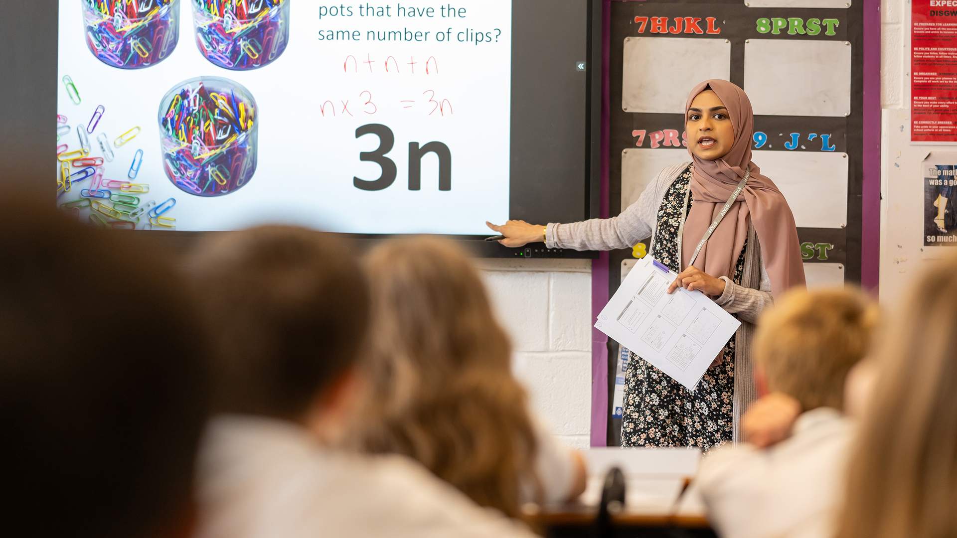A teacher stands in front of a class. She is pointing to a whiteboard with algebra equations written on it.