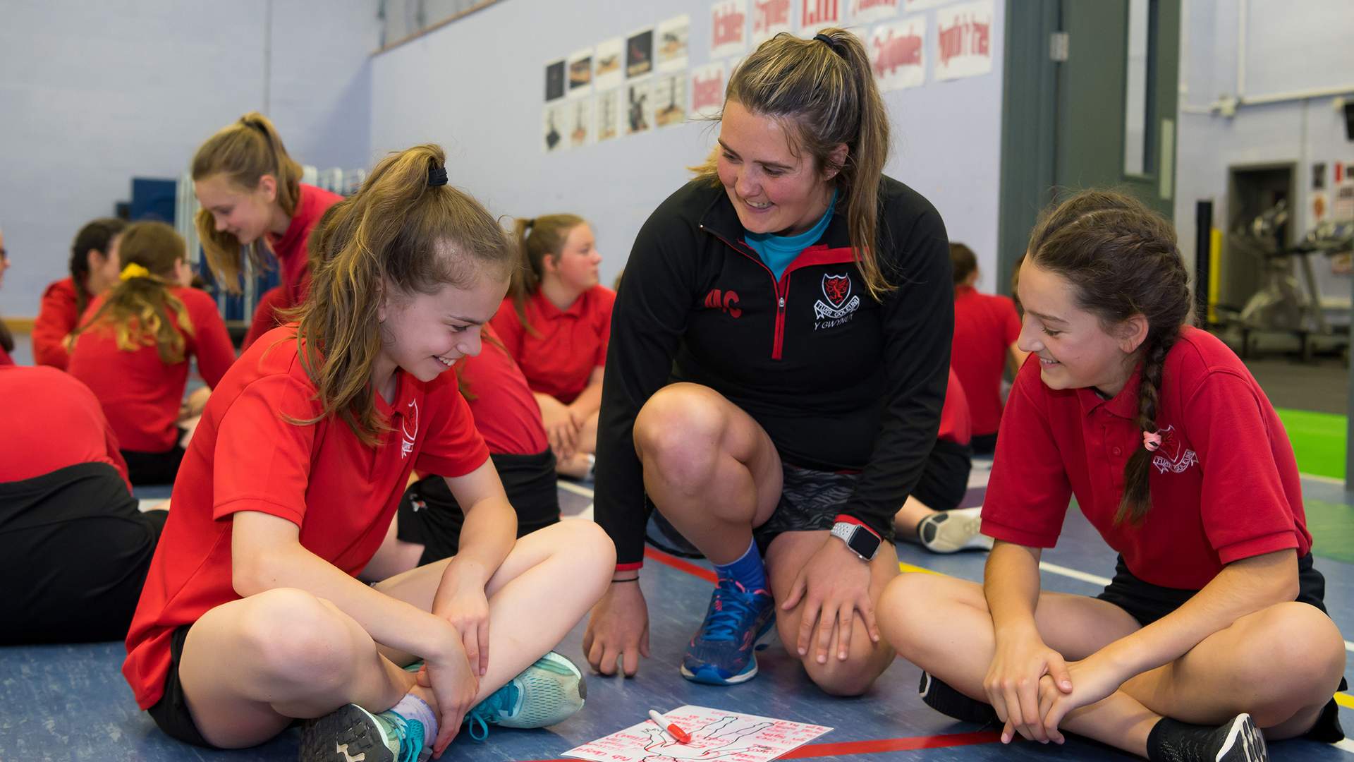 A teacher kneels between two pupils sitting on the floor of a gymnasium.