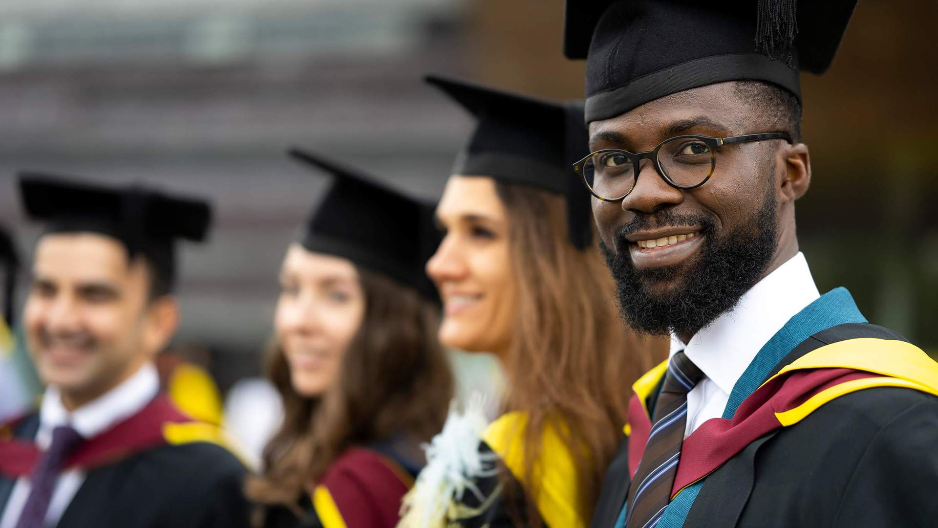 A graduate in cap and gown. In the background are three other graduates also wearing caps and gowns.