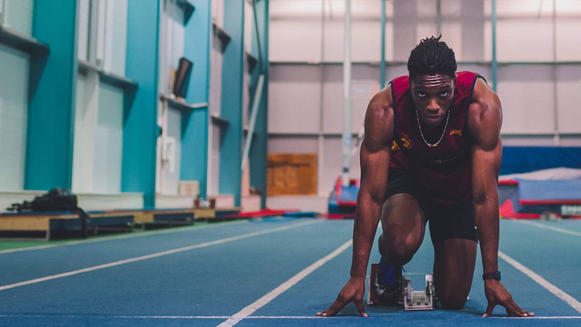 An athlete crouches in a starting position on a blue indoor track, preparing to sprint.