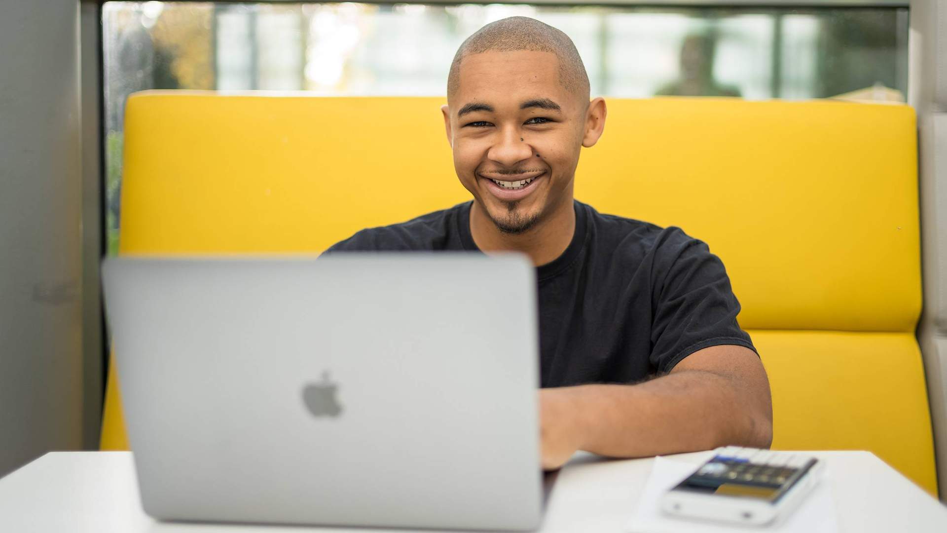A person is smiling while sitting at a table with a laptop in front of them. They are in a bright, modern setting, with a yellow cushioned seating area behind them.