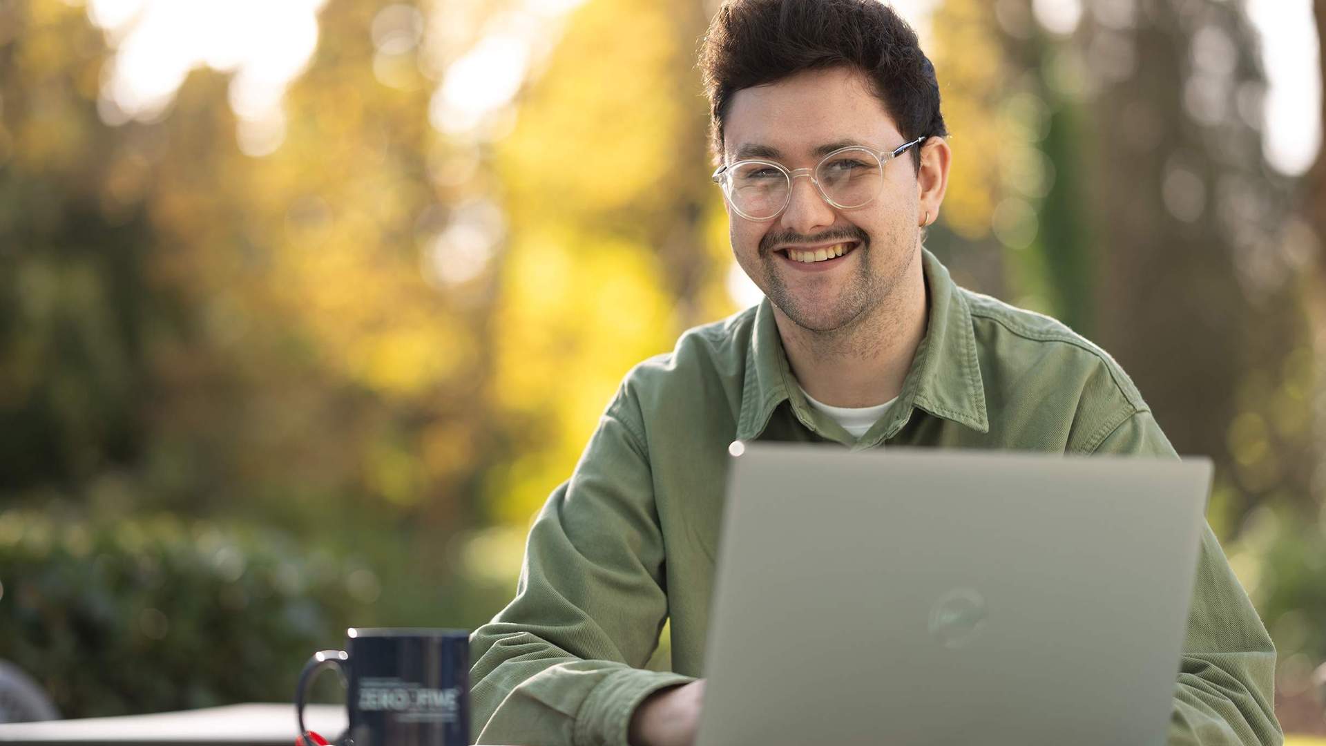 A young man sitting outside at a table. In front of him is a laptop computer and coffee mug.