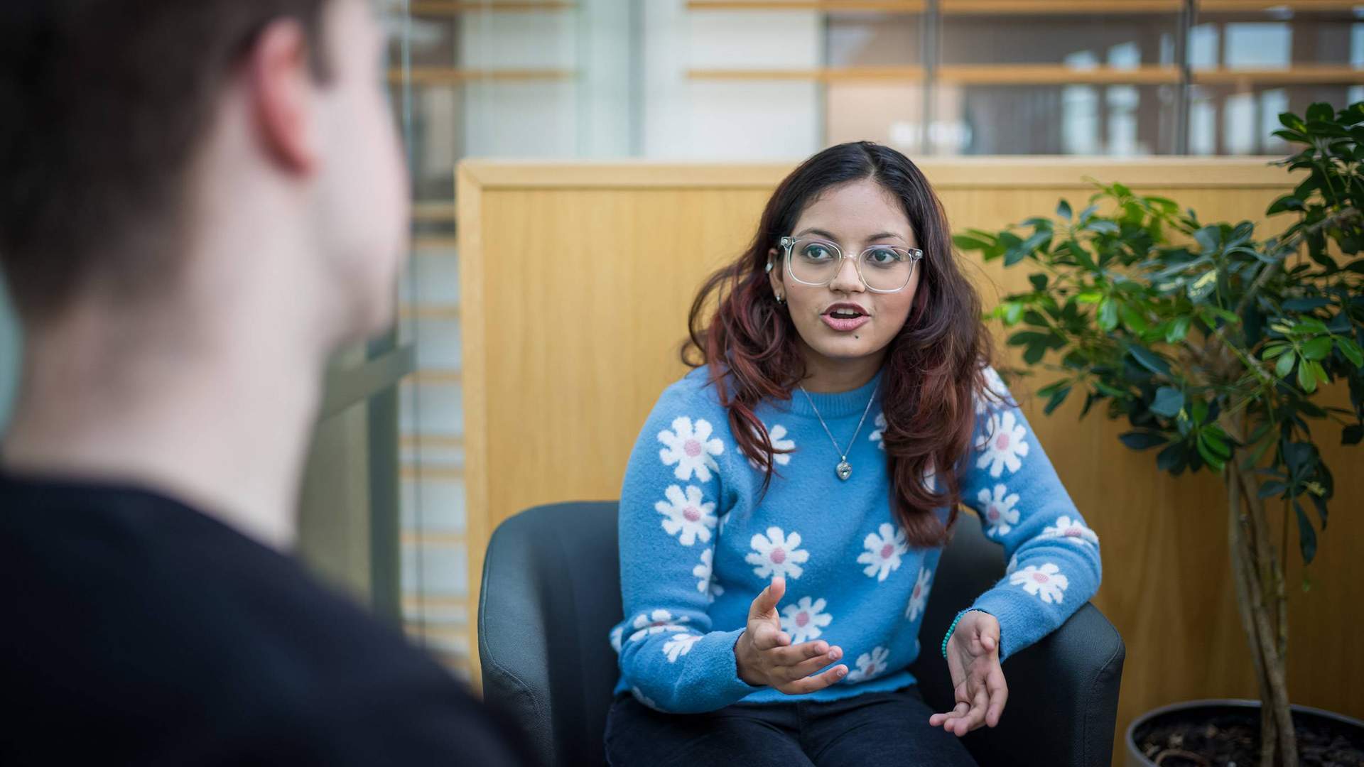 A young woman with glasses and long hair, wearing a blue sweater with white flower patterns, gestures while talking. She is seated in a modern indoor space, facing another person whose back is to the camera. A green plant is in the background.