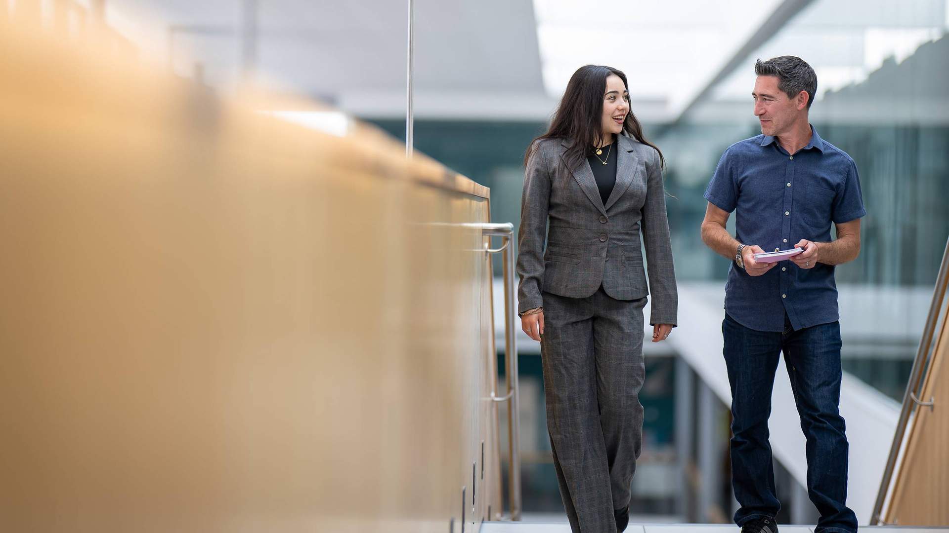 Two people walk alongside each other at the top of a staircase. In the background are glass walls overlooking an open atrium space.