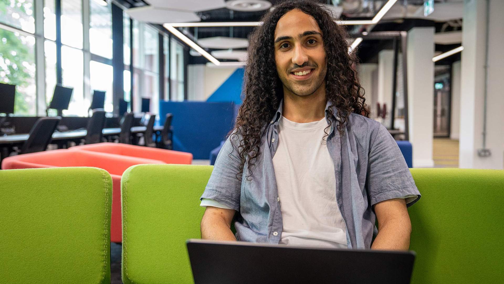 A young man sits on a green seating area with a laptop computer on his lap. In the background are rows of computer workstations.