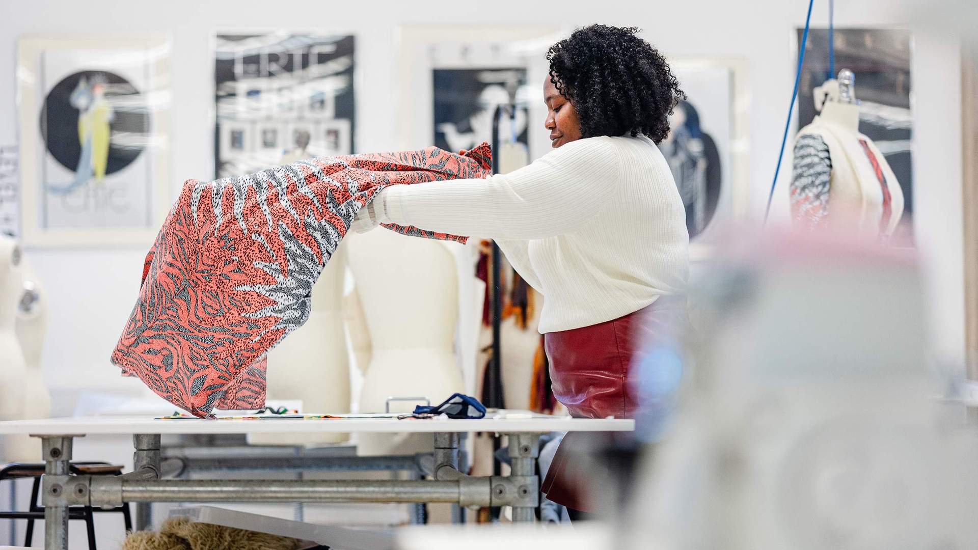 A person with curly hair holds up a colourful fabric in a design studio. In the background, a mannequin and framed art are visible. The room is well-lit, creating an inspiring workspace for creativity.