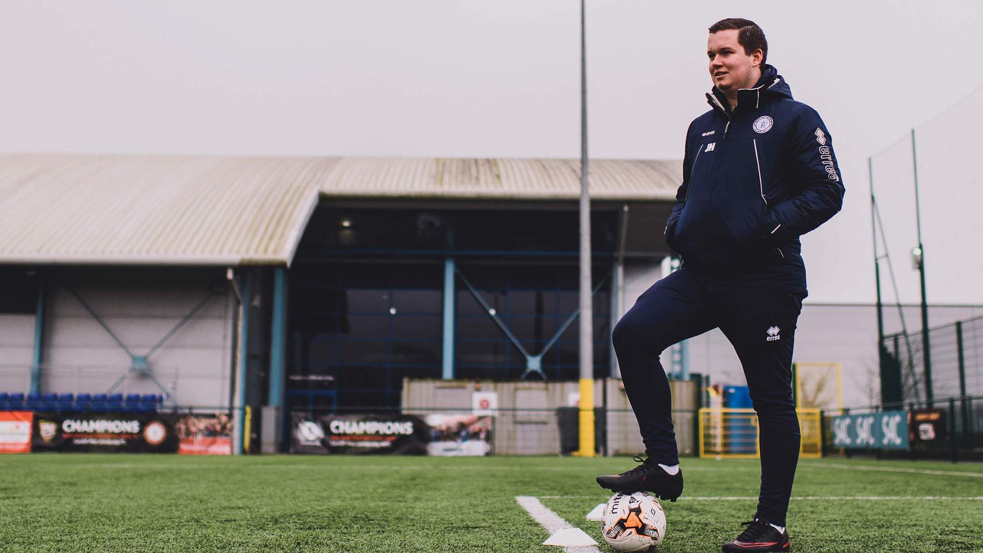 A football coach standing on the edge of the goal area with his foot on a ball.