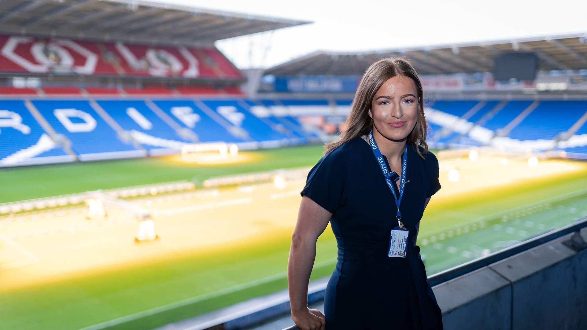 A woman in a stands smiling on the balcony of an empty stadium. The stadium seats are predominantly blue, with some red sections, and green grass covers the field. She wears an ID lanyard around her neck.